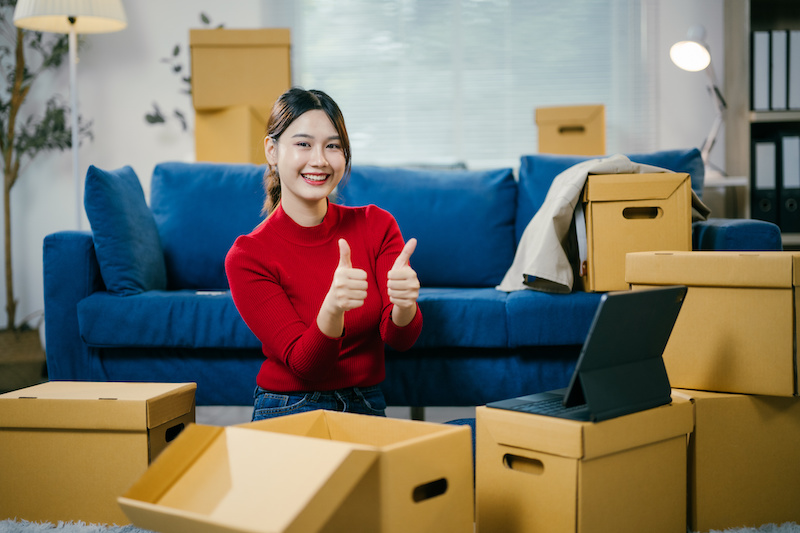 Young woman is smiling and giving thumbs up while unpacking boxes in her new apartment. She is happy with the service provided by the moving company