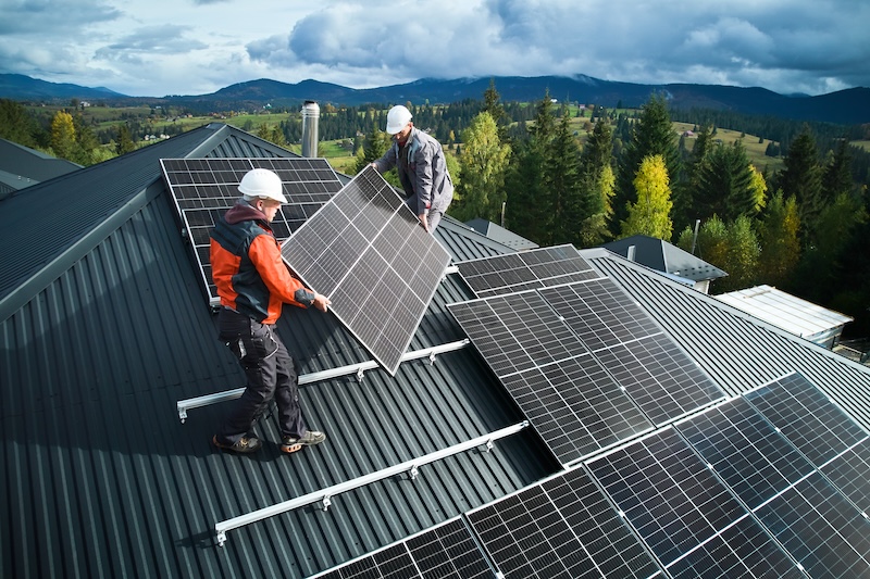 Workers building solar panel system on roof of house. Men technicians in helmets carrying photovoltaic solar module outdoors. Concept of alternative and renewable energy.