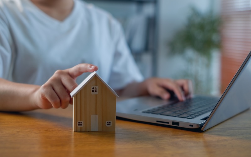 Person touching wooden house model beside laptop on desk, real estate and home finance concept for online property search, mortgage planning, housing investment and smart decision making.