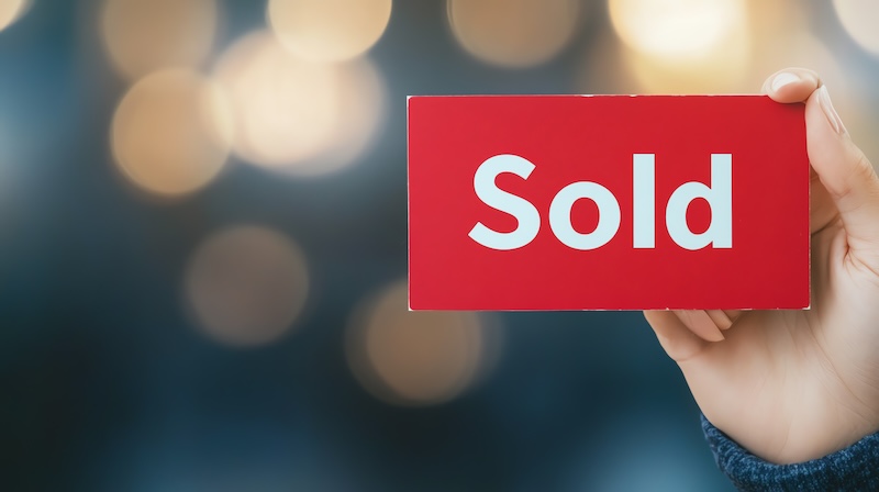 A hand holding a red 'Sold' sign against a blurred background, symbolizing a successful sale or transaction in a vibrant market.