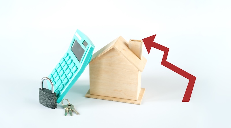 A conceptual still life featuring a wooden house model, a teal-colored calculator, a black padlock, and a set of keys on a plain white background. A large red upward-pointing arrow indicates an increase.